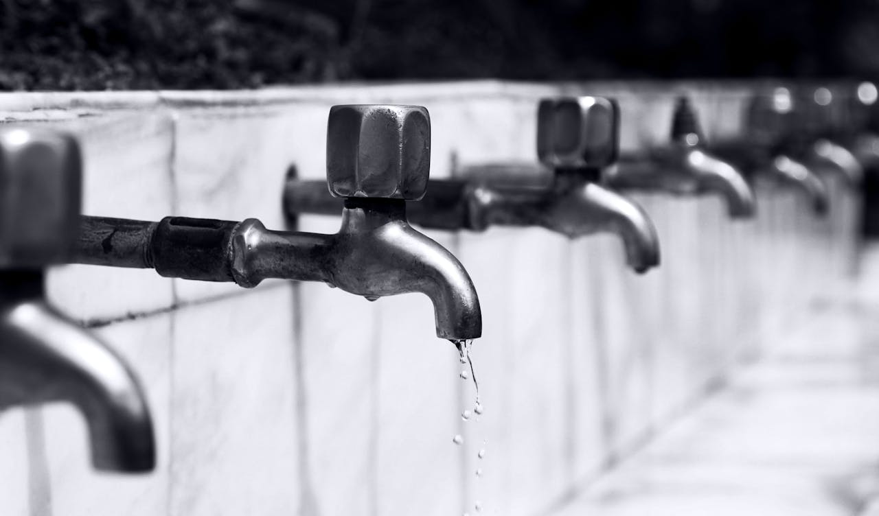 Black and white image of a row of faucets with water dripping, highlighting plumbing details.